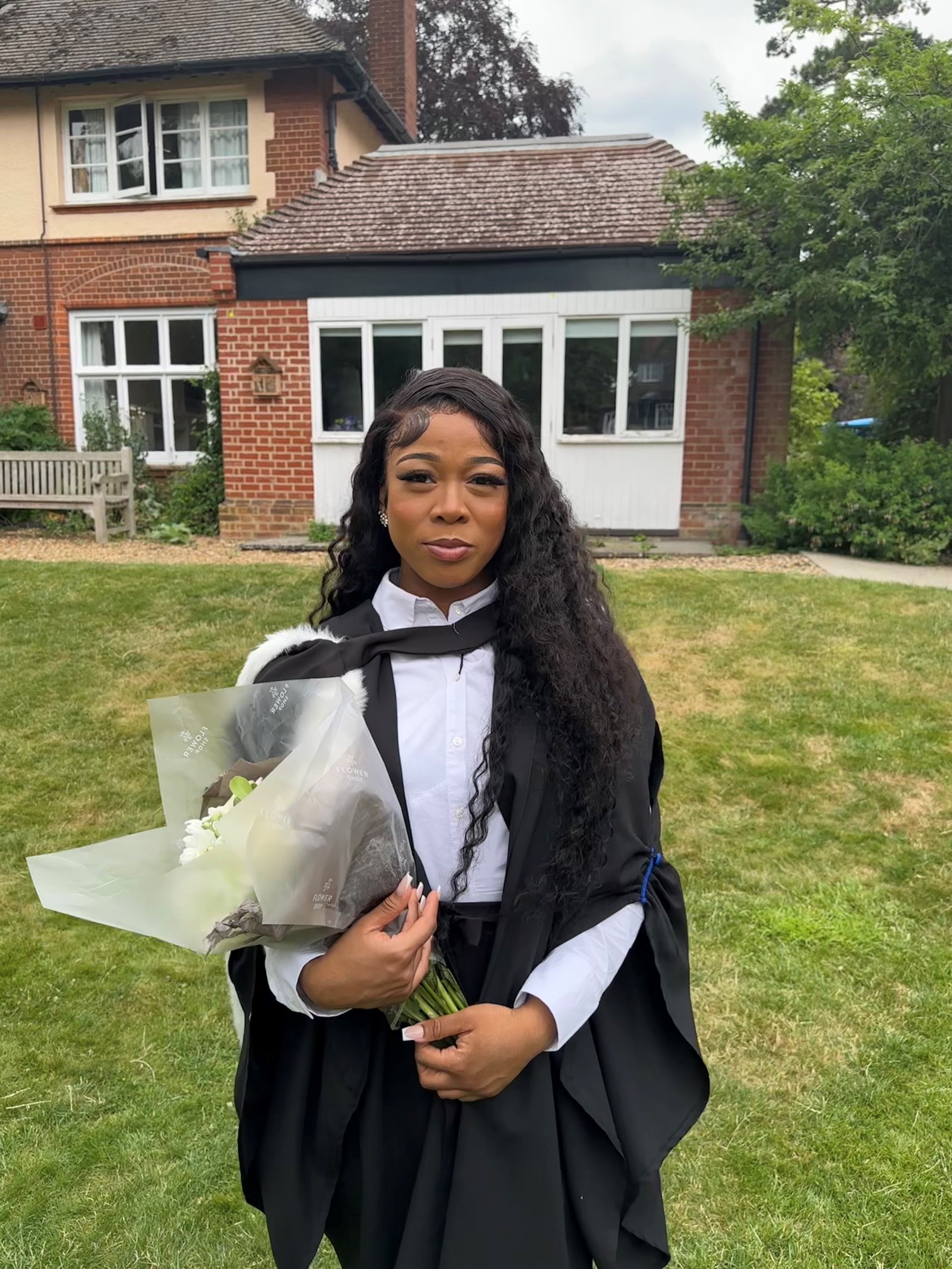 An image of a Black woman holding flowers and wearing graduation robes in a garden