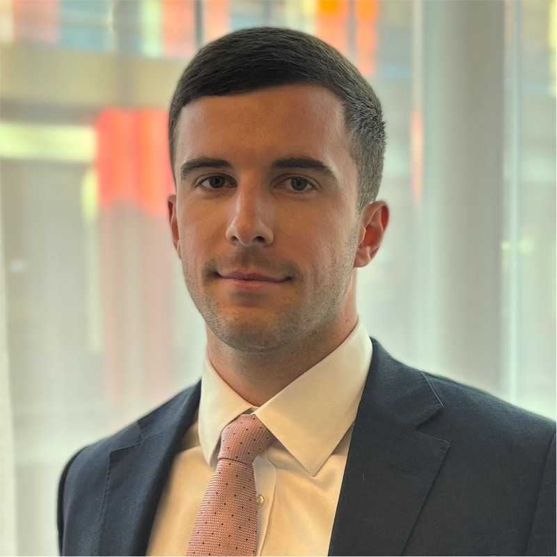 An image of a man with short hair brown hair in an office environment wearing a blue suit and pink tie