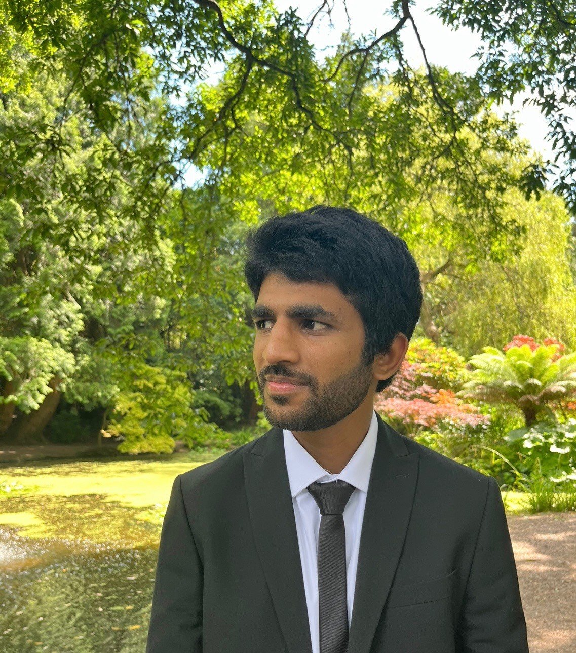A headshot of a man wearing a black suit and tie in a garden setting