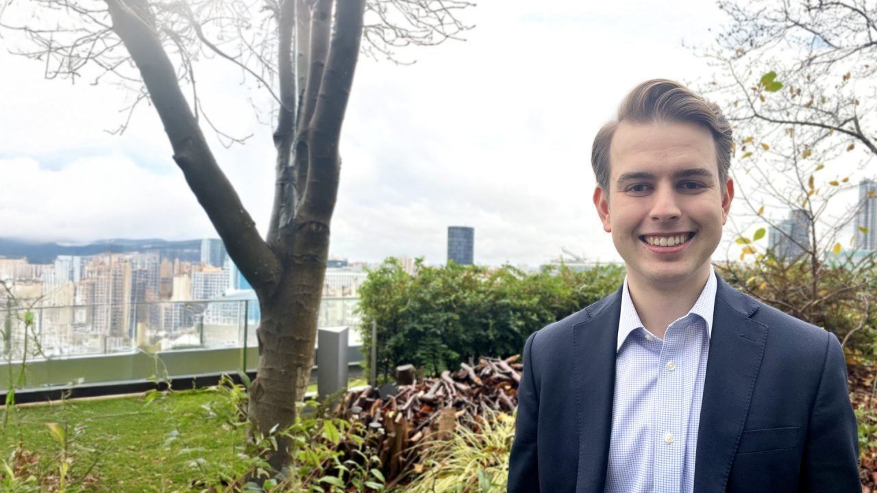 A man with brown hair, wearing a blue suit standing in a garden overlooking a city