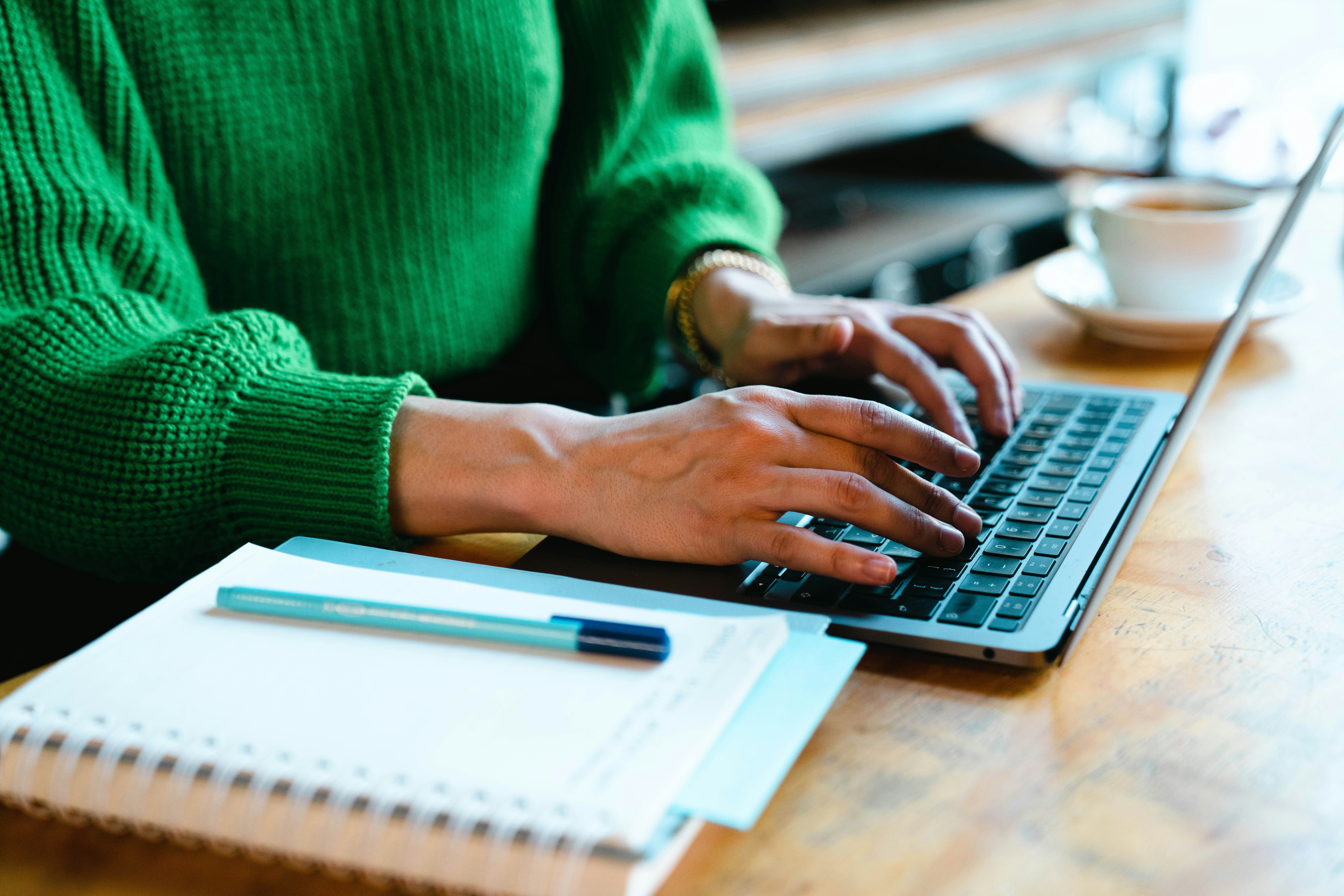 Woman typing on a laptop. Next to her is a notebook and a pen.
