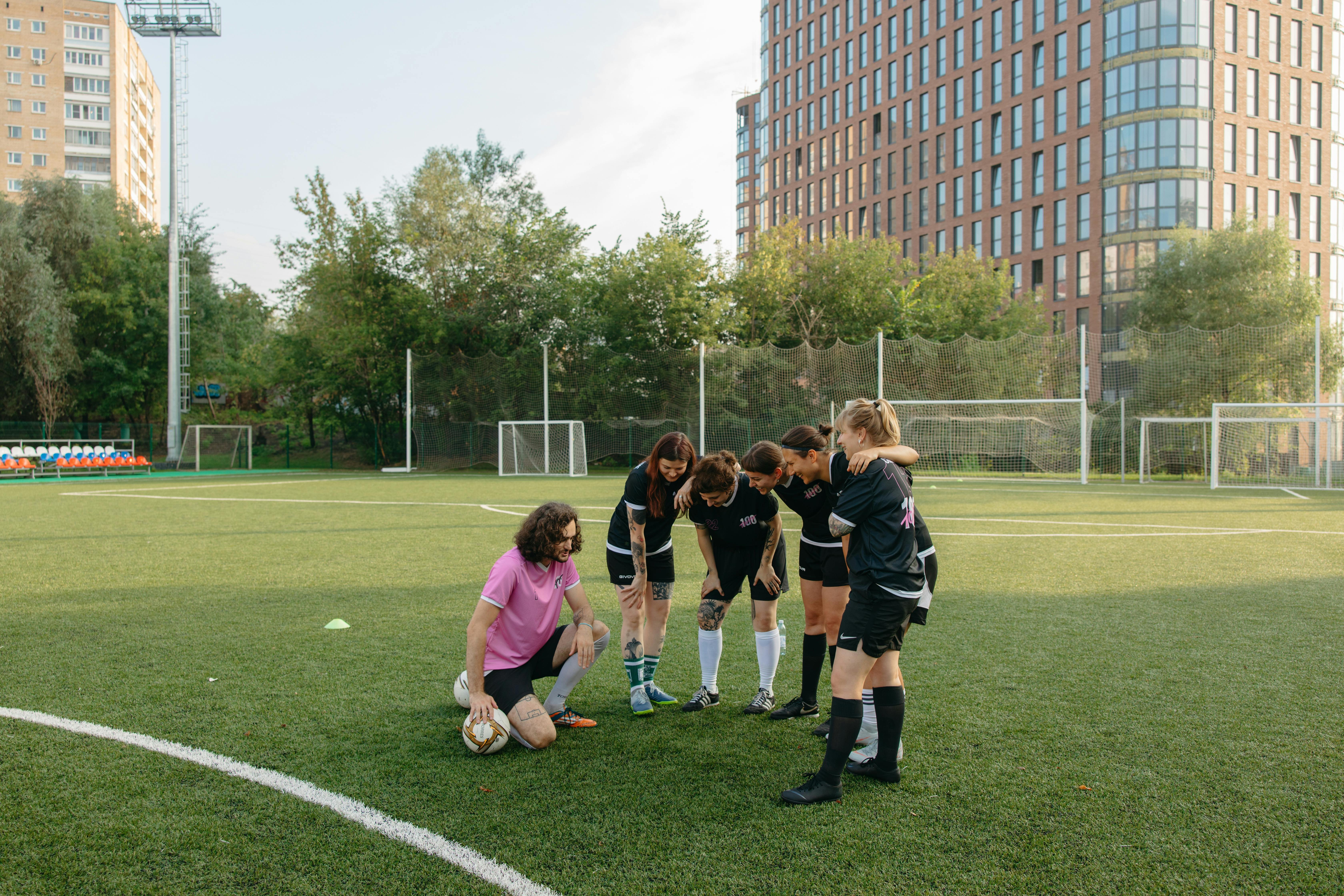 A team of five women stood in a huddle on a football pitch beside their male coach, who is crouched down on the ground.