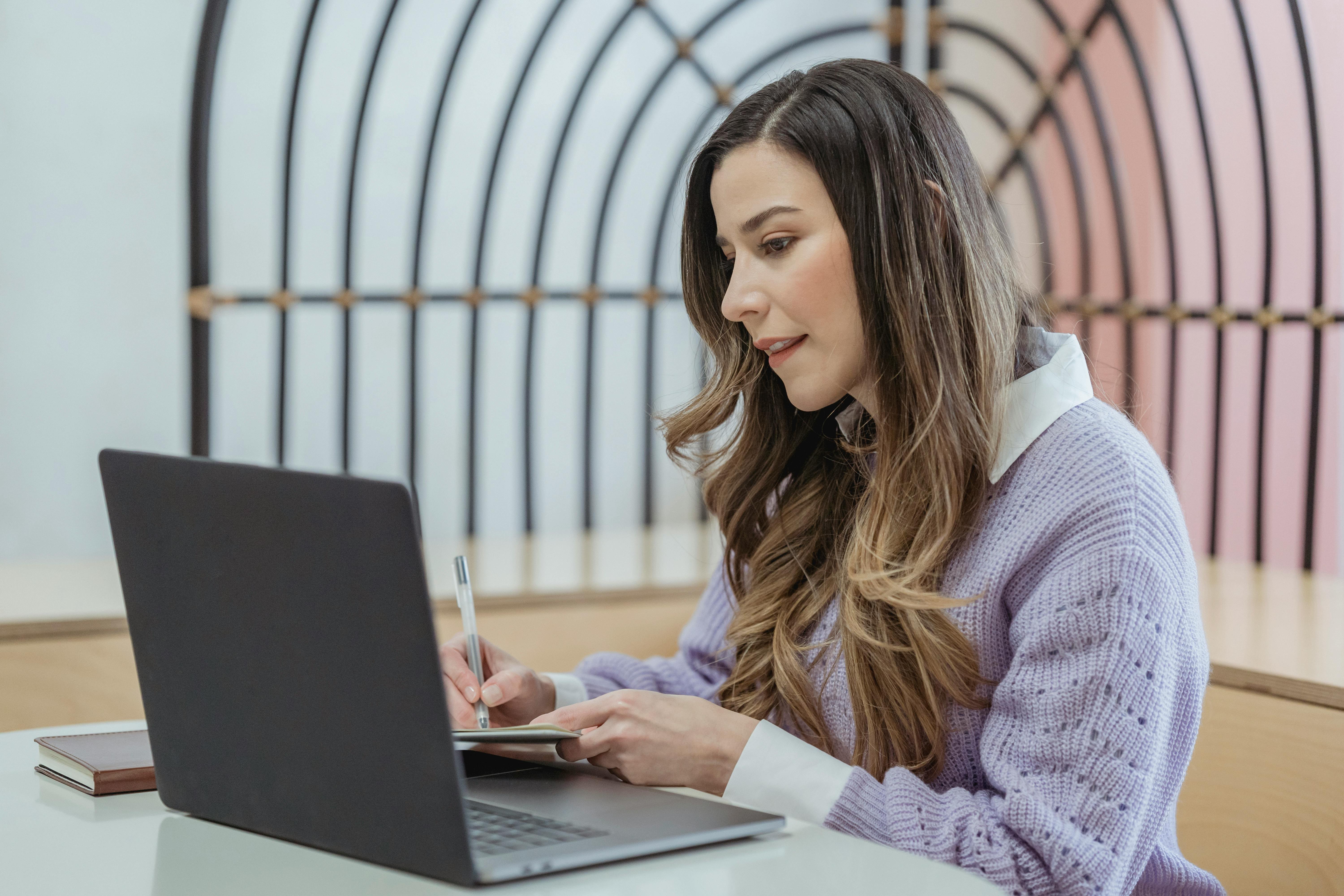 Woman looking at a laptop and taking notes down in a notebook. She is wearing a lilac jumper over a white shirt.