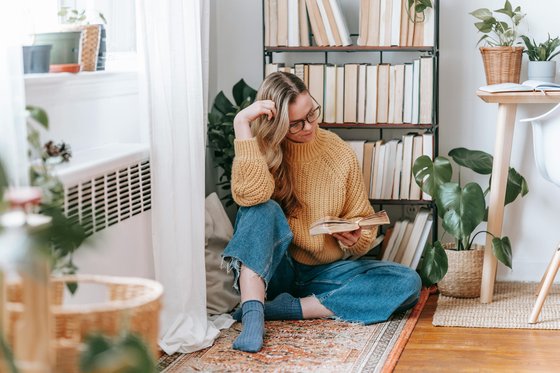 A woman in denim jeans and a yellow jumper sitting on the floor near a bookshelf while reading a book
