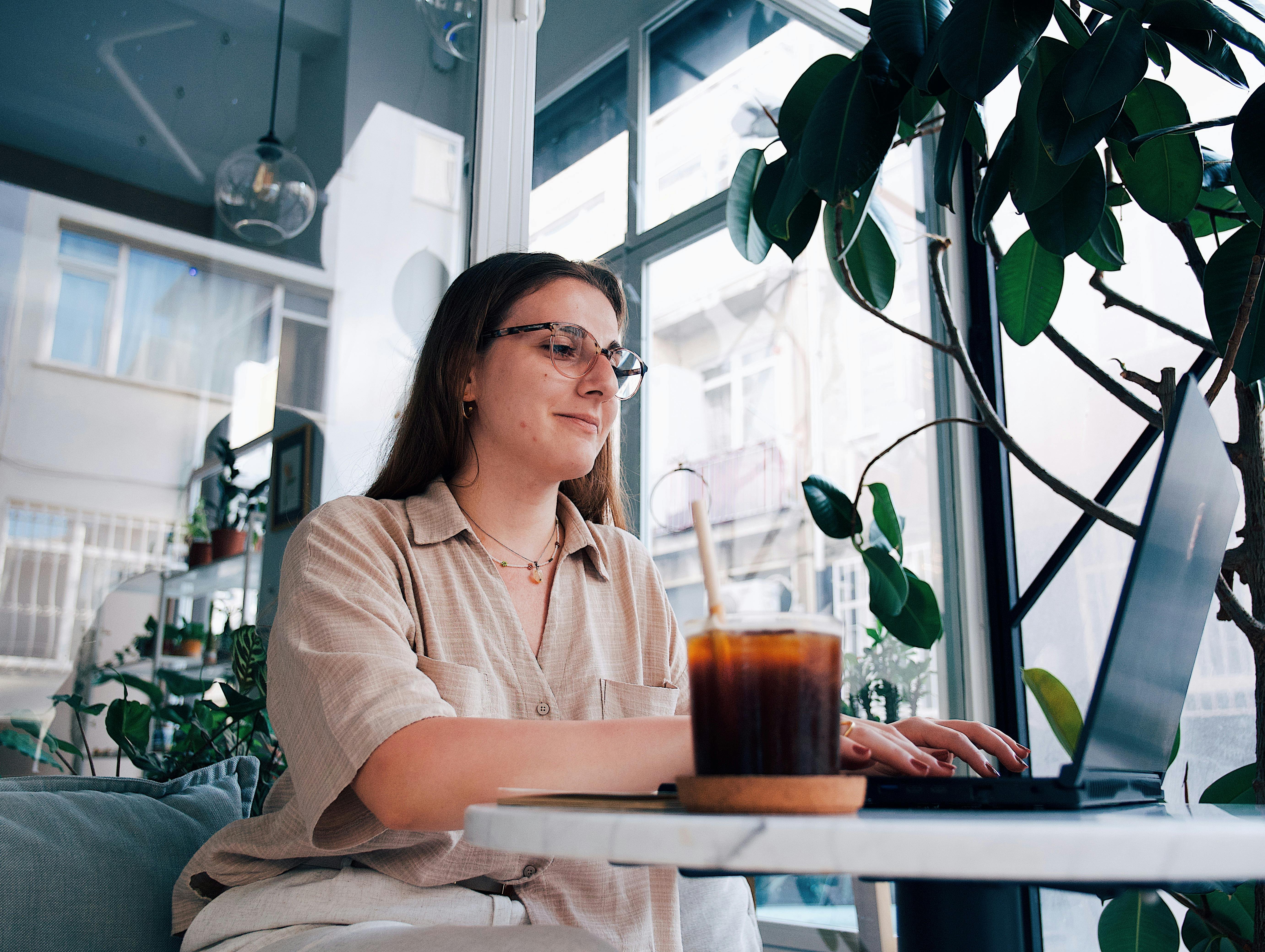 A woman sat at a table using a laptop. She has a cold brew coffee next to her and lots of houseplants in the room.
