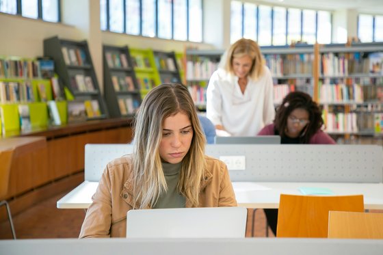 Young woman sat in a library on her laptop. In the background, there are other students on laptops and a member of staff is helping them.