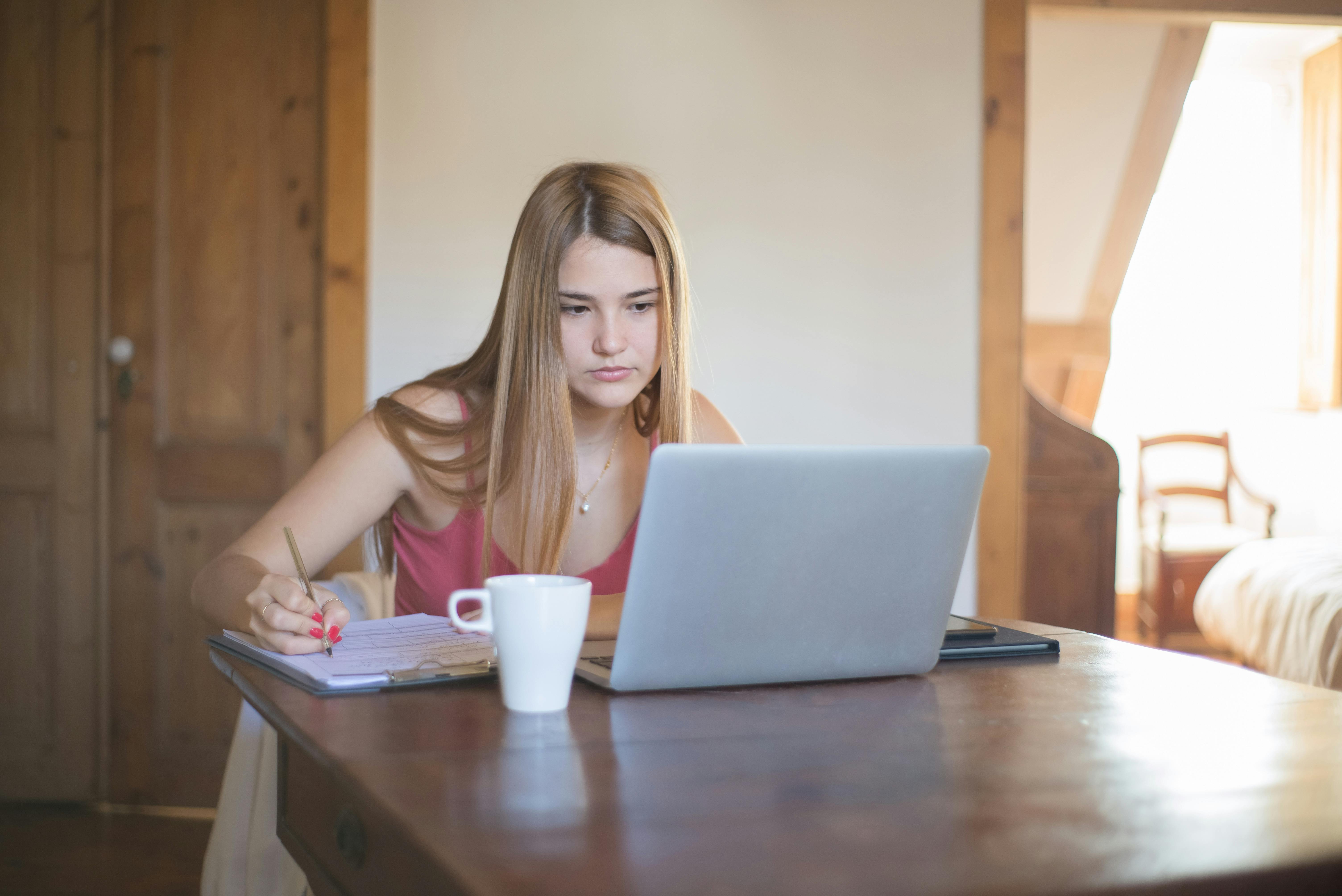A young woman using a laptop and writing down notes.