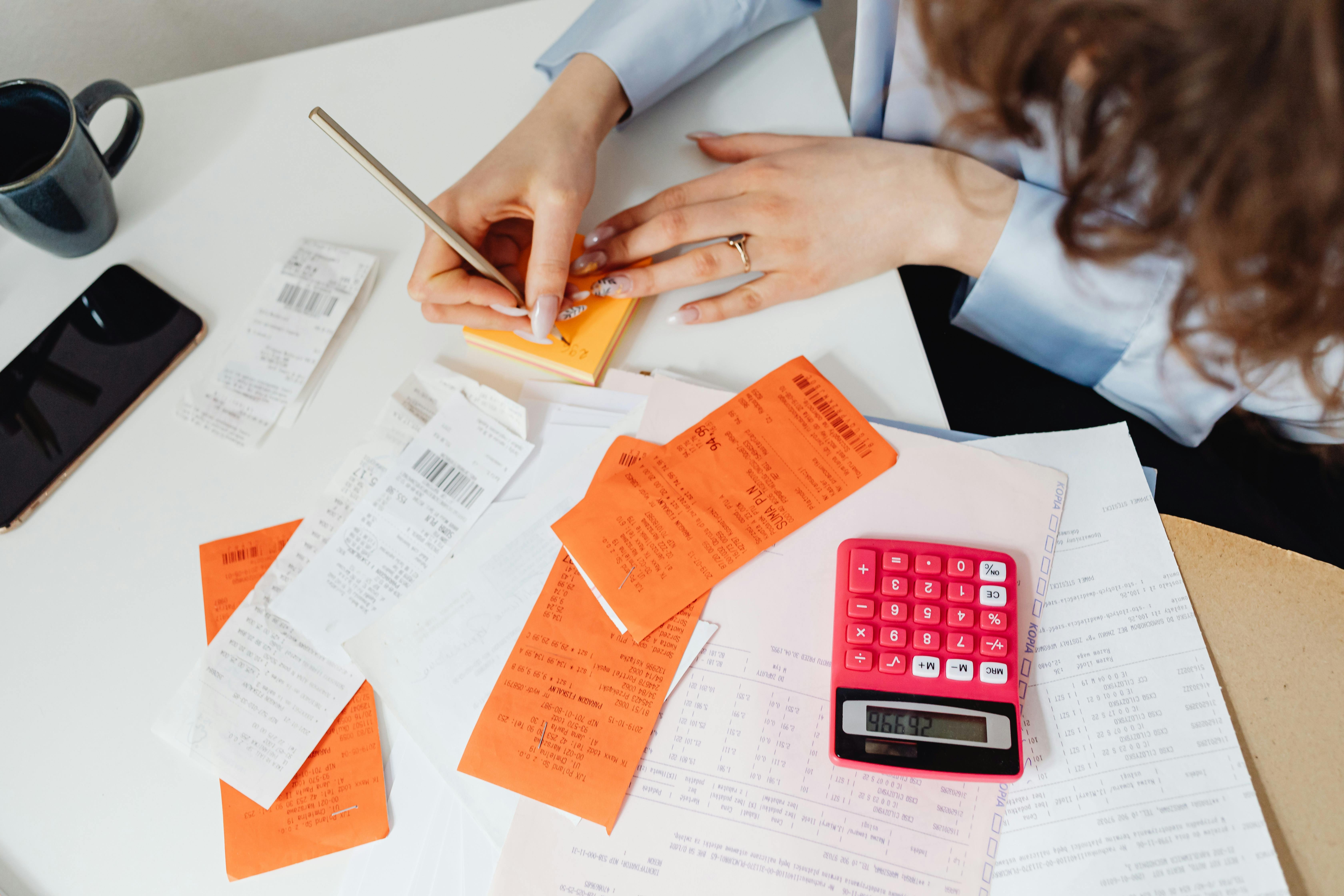 An aerial view of a woman surrounded by receipts and bills. She has a calculator and is writing something down on a sticky note.