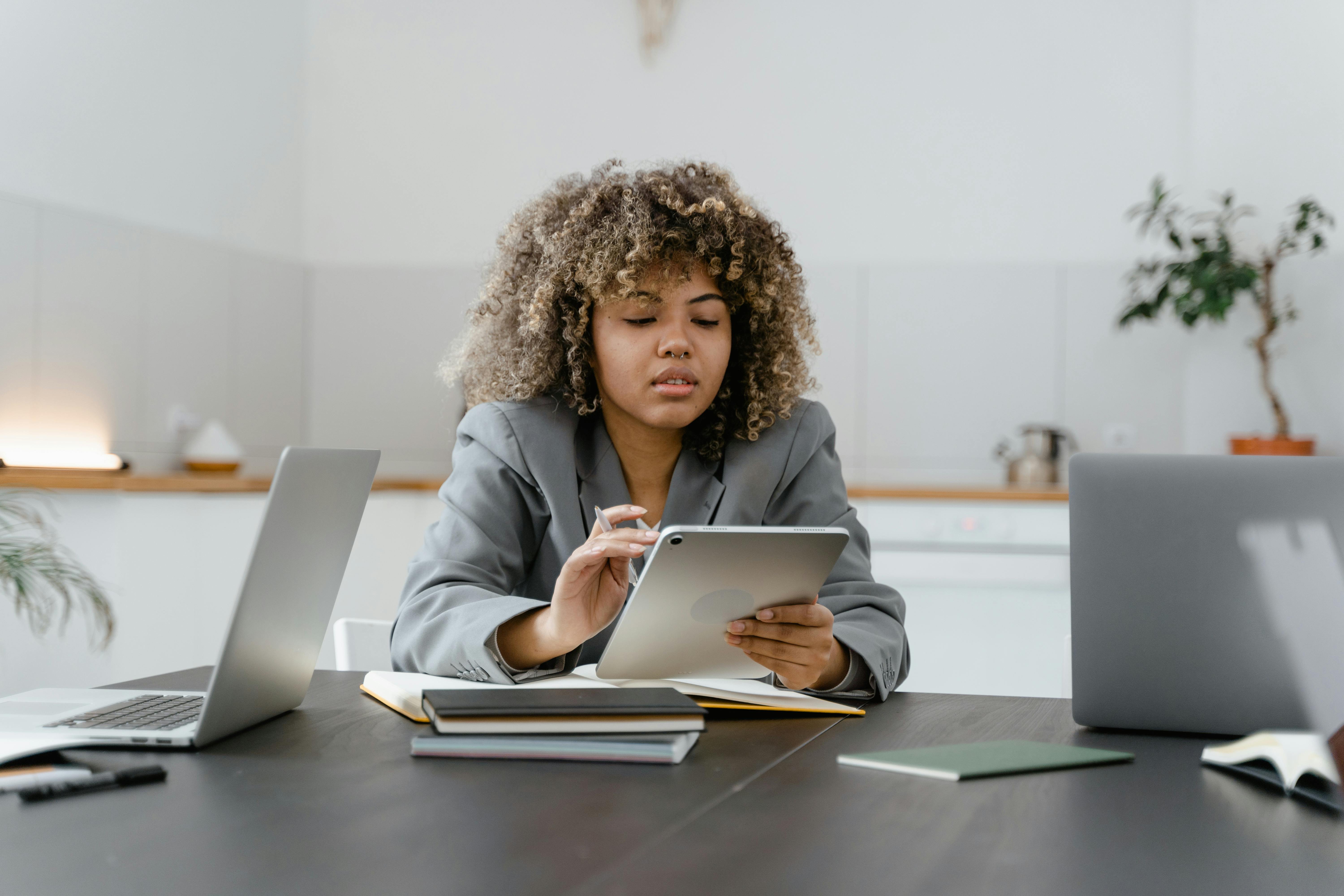 Woman sat a table looking at a tablet device. She is surrounded by several notebooks and two laptops.