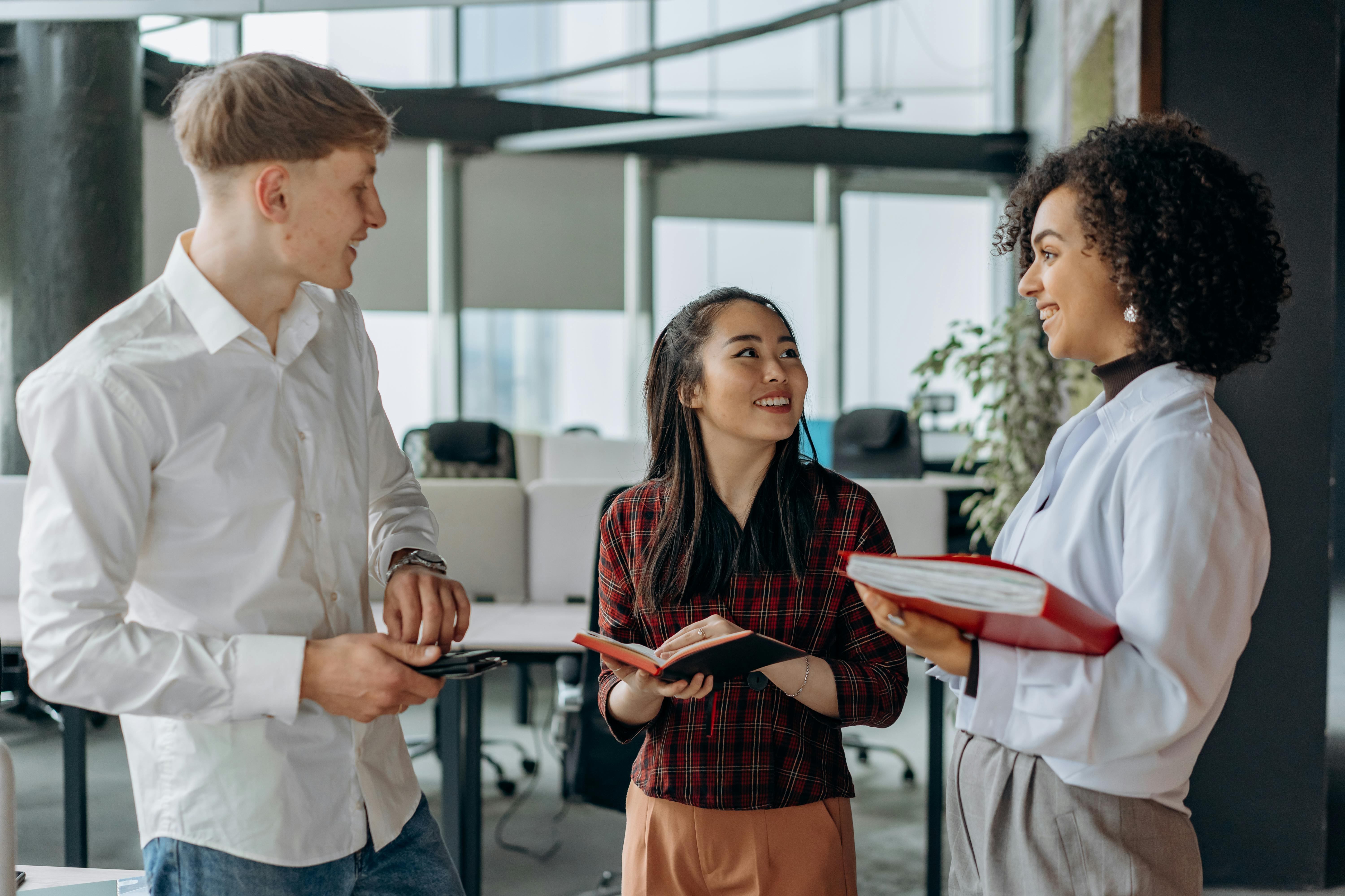 A man and two women standing in an office environment smiling. The man is holding a phone, one woman has an open book and the other woman is carrying a red folder.