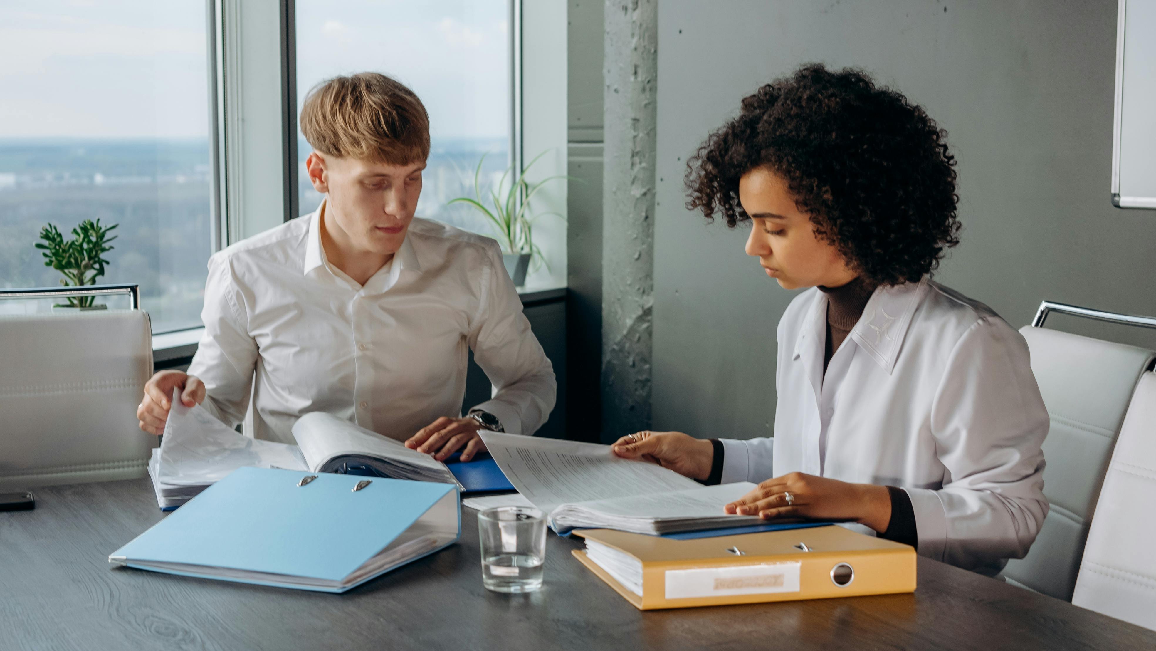 A man and a woman, both wearing white long-sleeved shirts, sitting and reading financial reports in a meeting room.