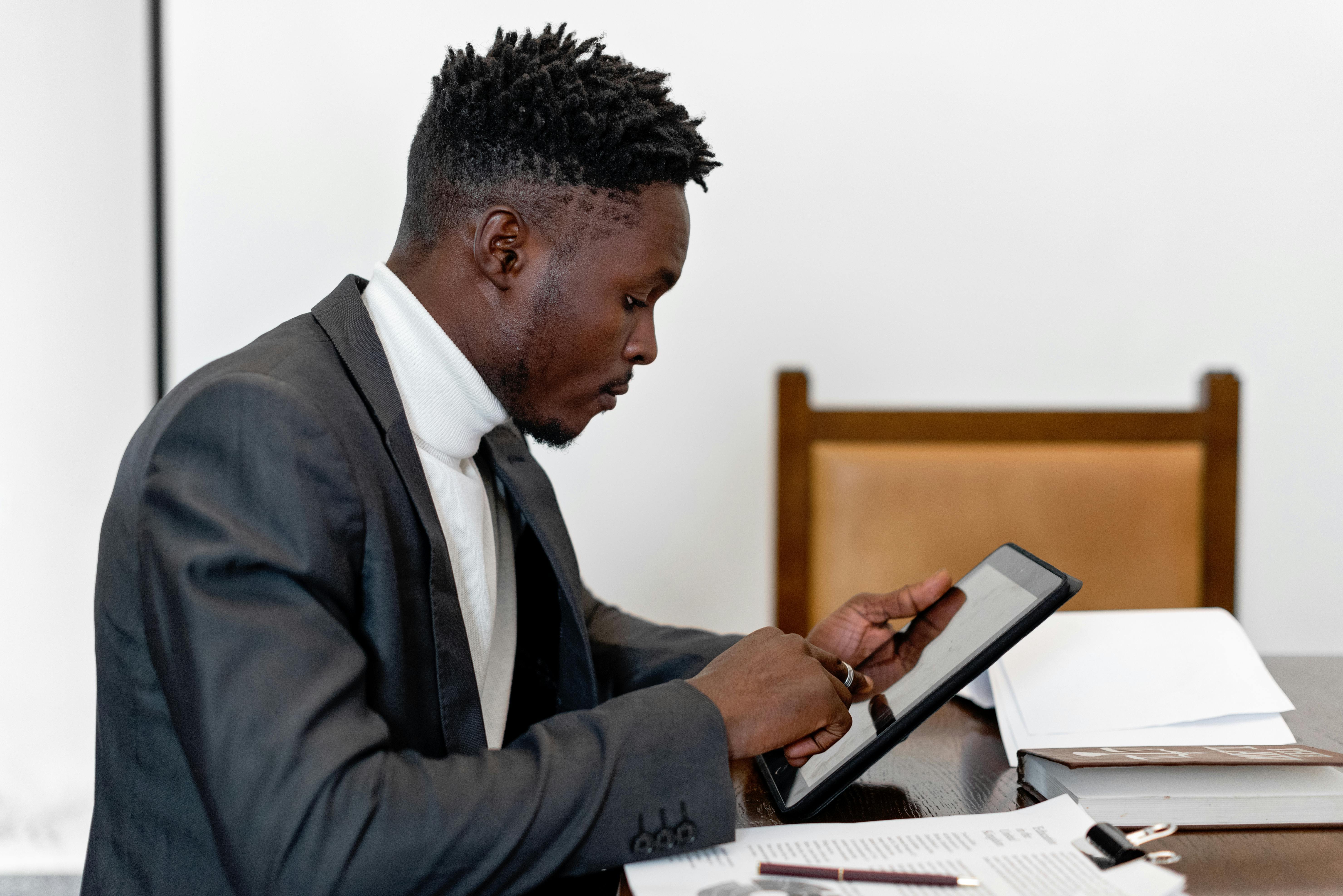 Smartly dressed man sat at a table looking at a tablet while surrounded by paperwork.