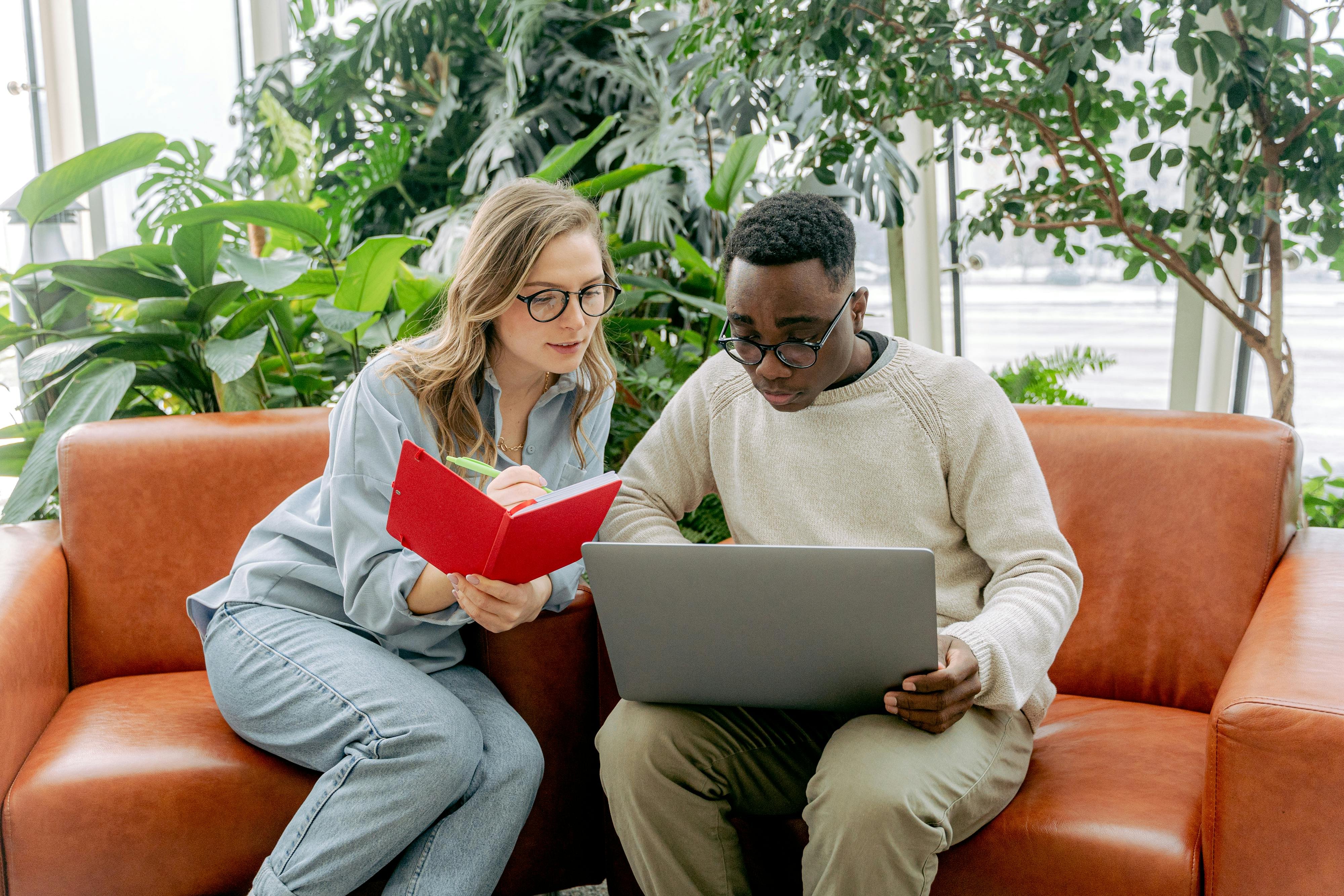A man and a woman sat on two leather chairs in an office environment, looking at the man's laptop together. The woman is also taking notes in a notebook.