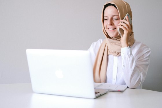 A woman smiling and sitting down while talking on her mobile phone. She is wearing a white smart shirt and a beige hijab. In front of her, there is a laptop and notebook on the table.