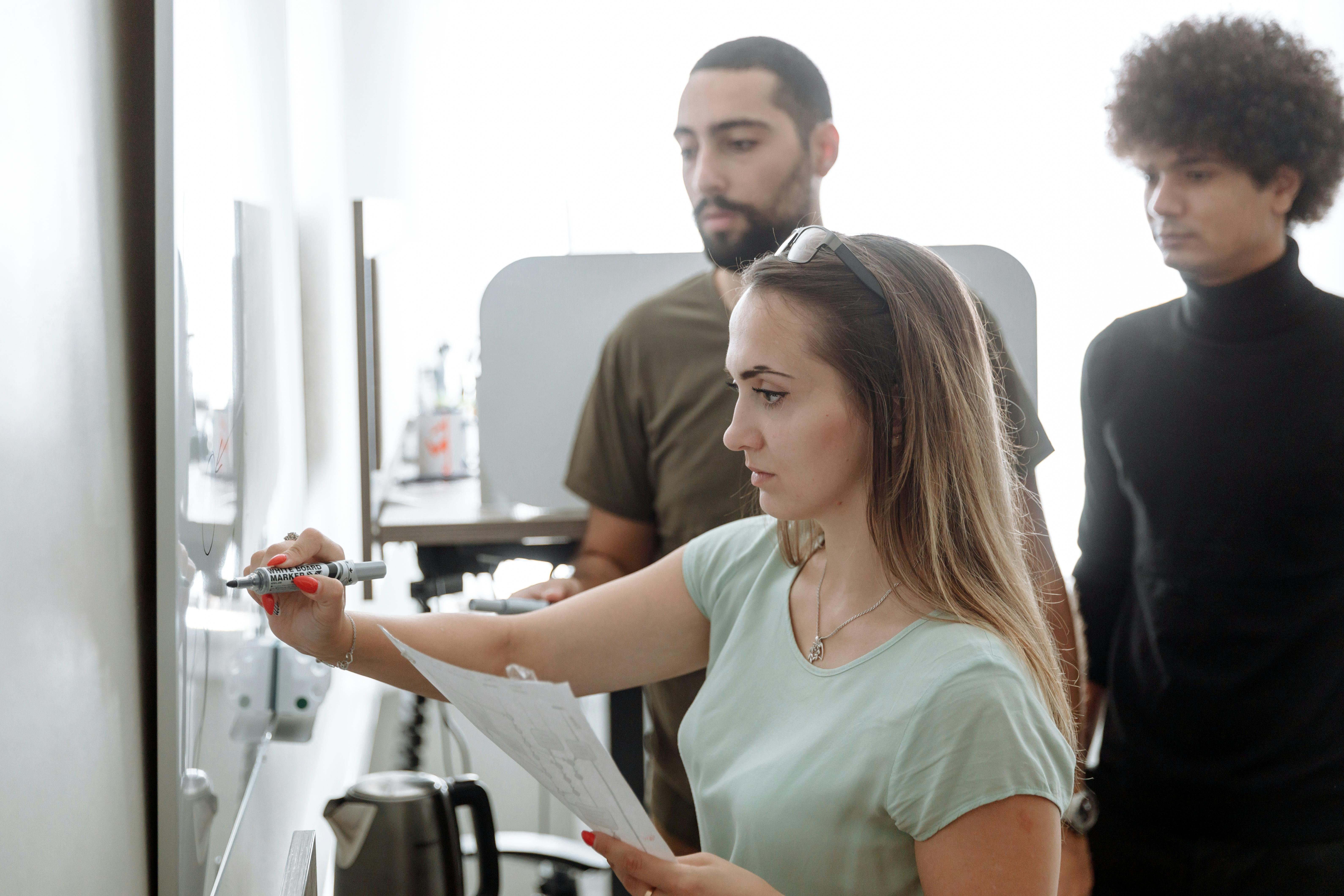 Three colleagues looking a large whiteboard. The woman at the front is writing on it, while holding a piece of paper.