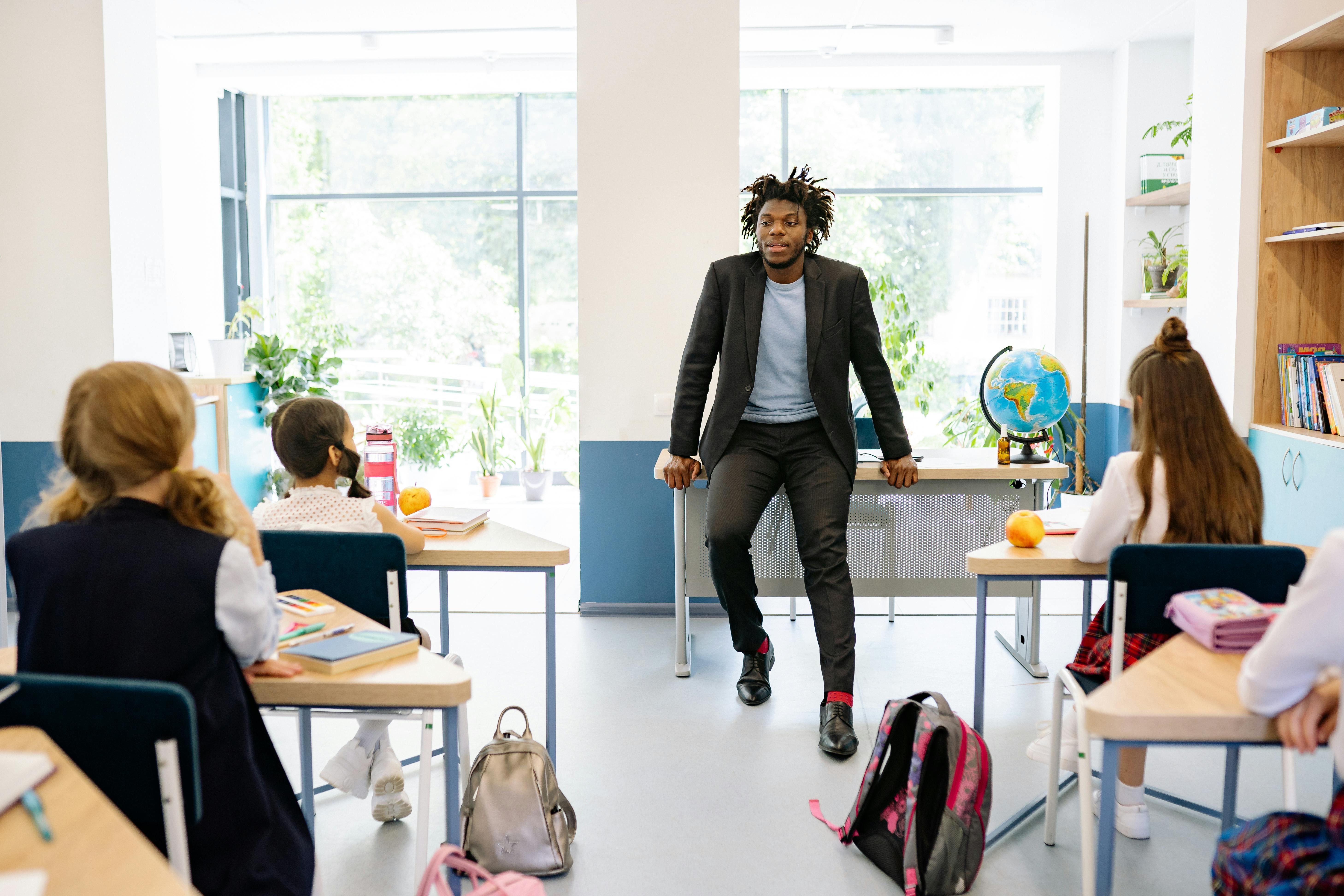 A male teacher stood in front of a classroom with children sat at individual desks.